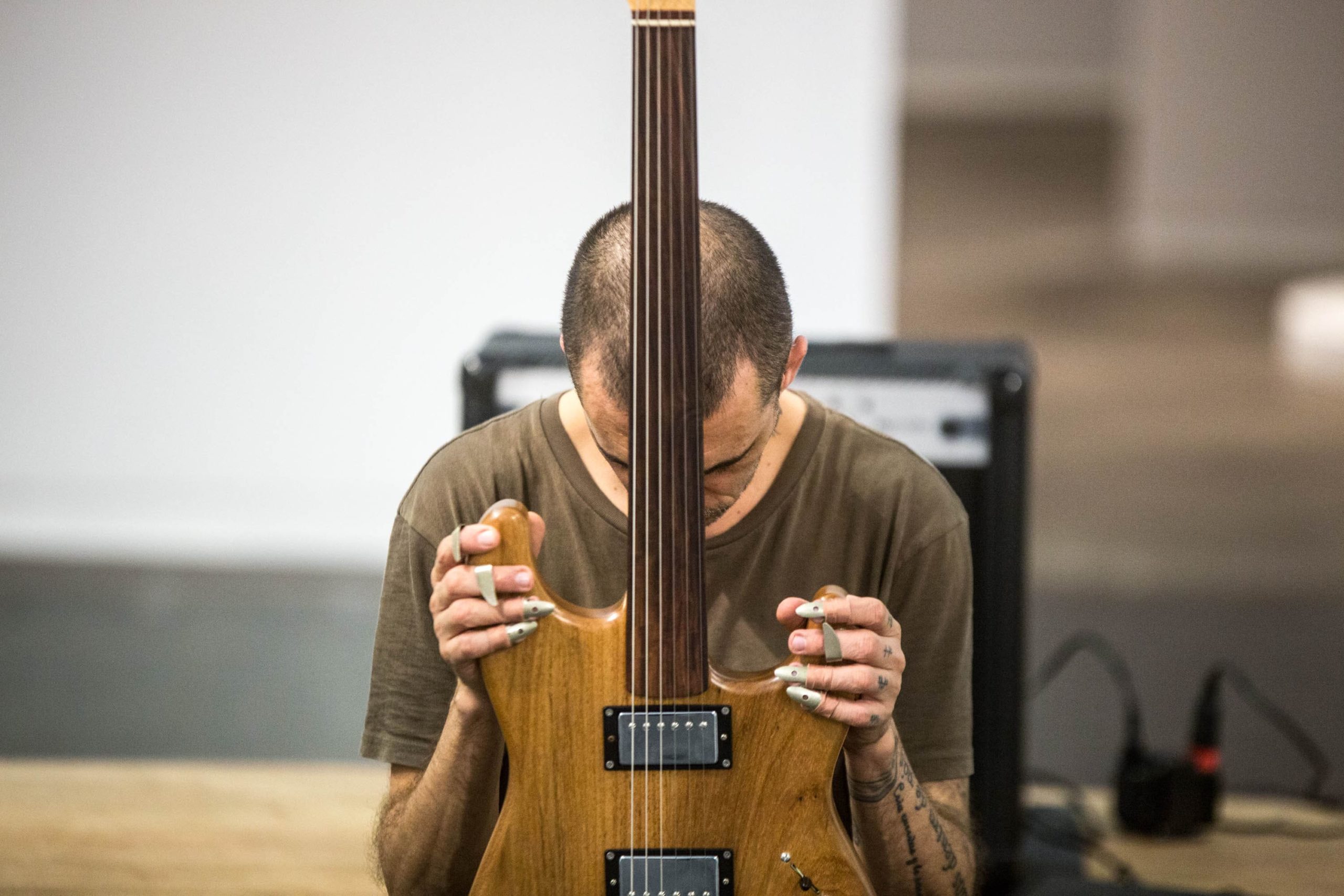 Alejandro Ahmed está no centro, de cabeça baixa, segurando uma guitarra à frente com as duas mãos.