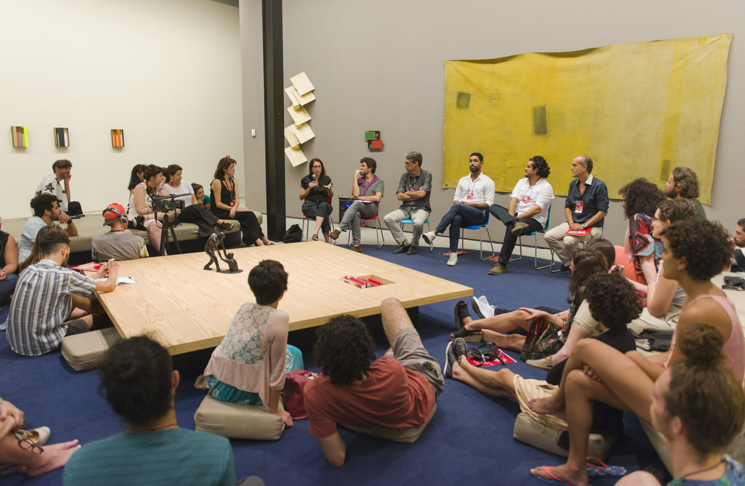 Imagem mostra pessoas sentadas reunidas em torno de uma grande mesa quadrada.